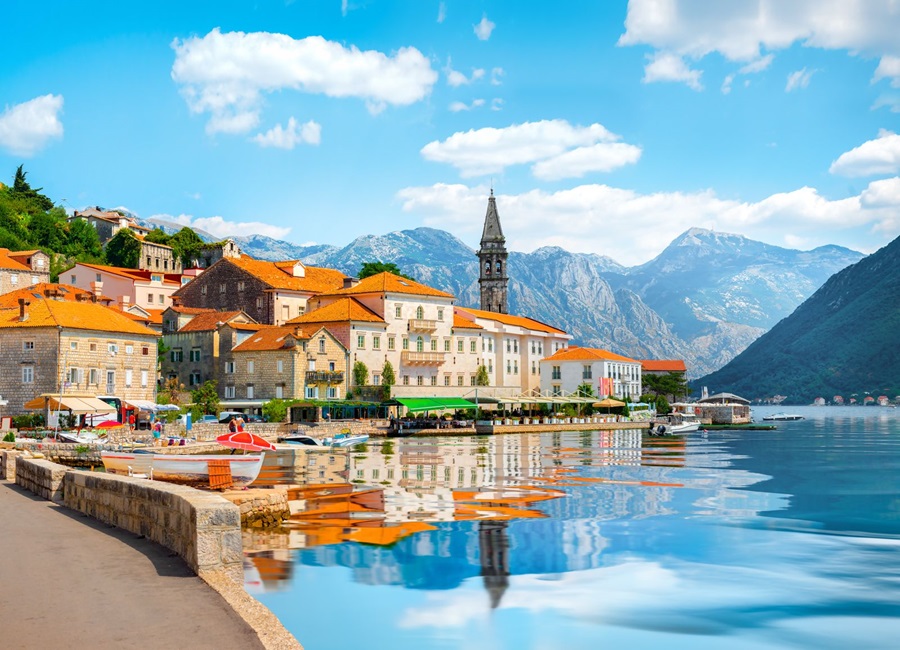 Coastal town with orange-roofed stone buildings along a calm harbor and a church bell tower, mountains in the background, reflections on the water.