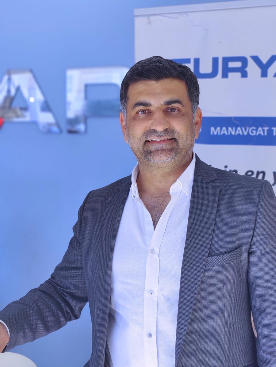 Middle-aged man in a gray blazer and white shirt posing for a photo in front of a blue backdrop with a sign in the background, smiling slightly.