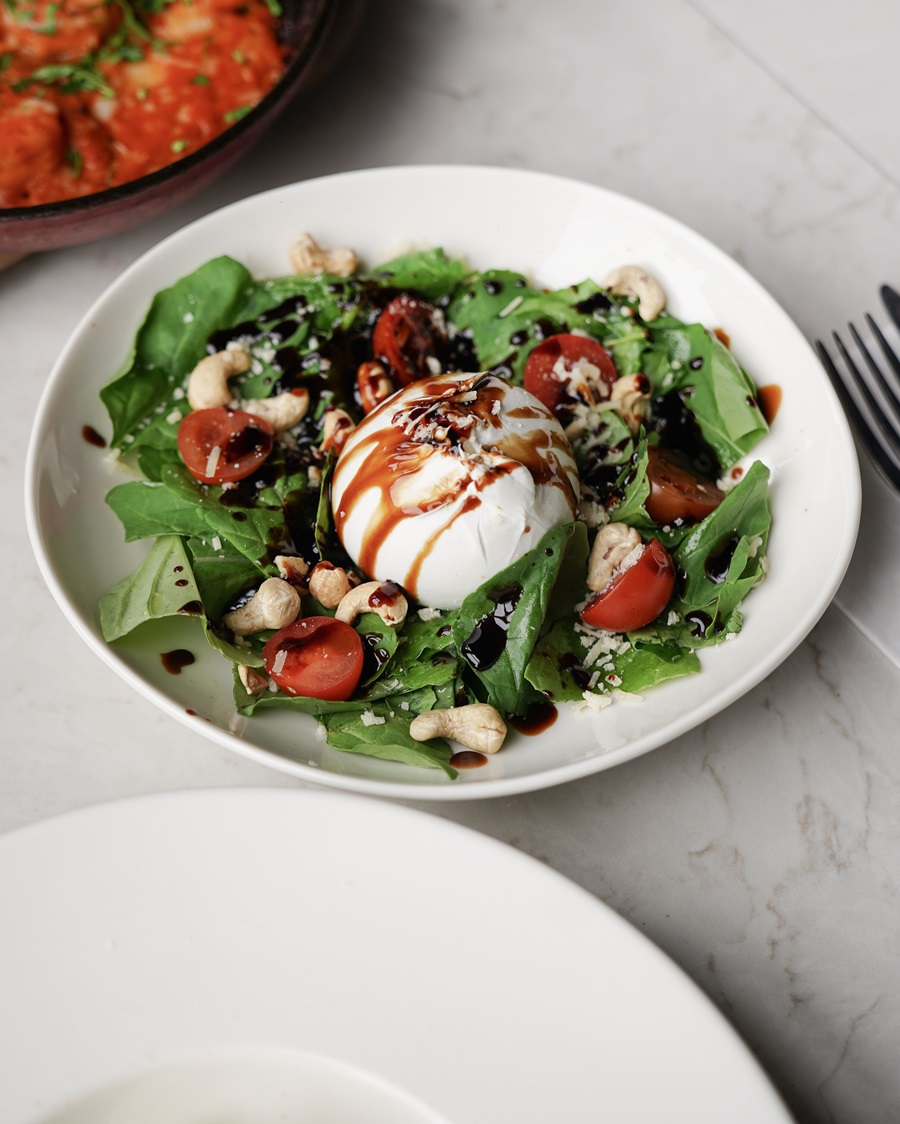 Burrata cheese on a greens salad with cherry tomatoes, cashews, and balsamic glaze drizzle.