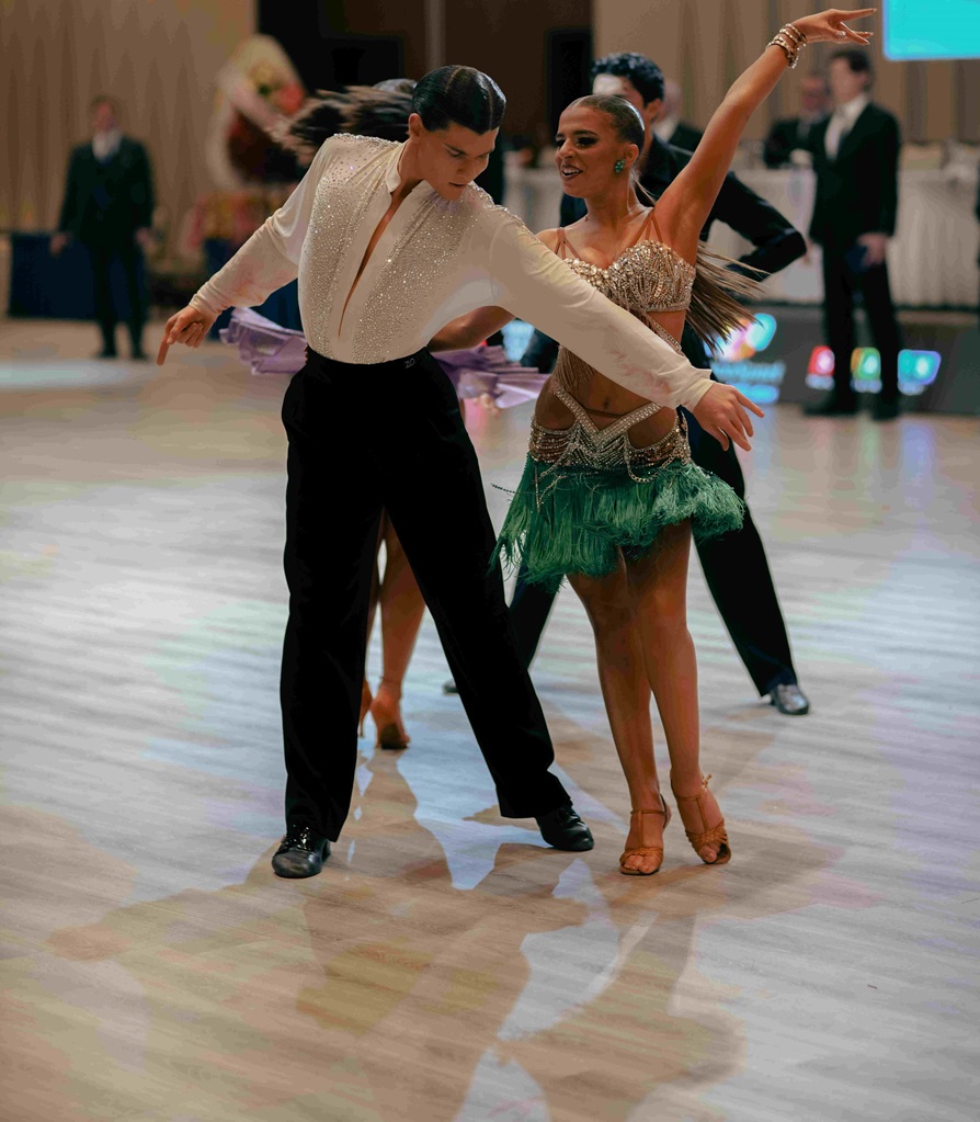 Ballroom dancers perform a dramatic dip on a polished competition floor; man in a white sequined shirt and black pants, woman in a green fringe dress.