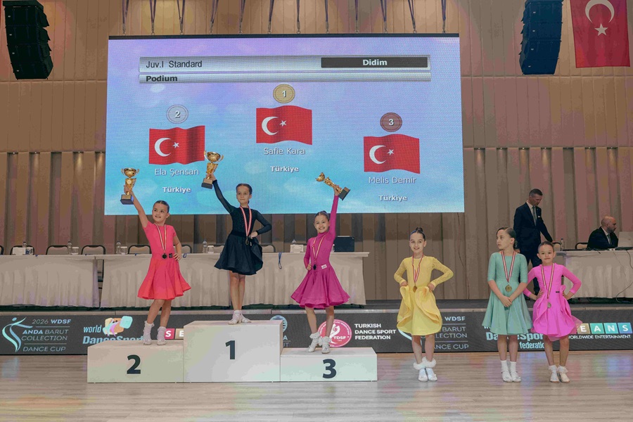 Young dancers on a podium hold trophies after a dance competition; large screen shows winners and Turkish flags in the background.