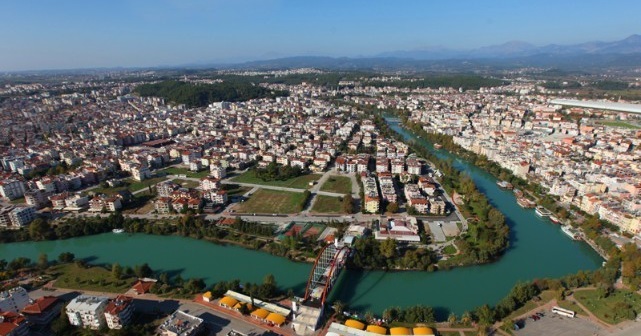 Aerial view of a town with a winding turquoise river, a red-arched bridge, and dense buildings surrounded by green hills in the distance.
