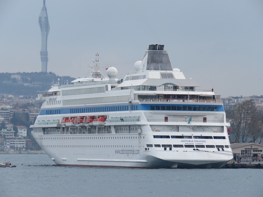 White cruise ship Astoria Grande docked at a harbor with a city skyline and a tall tower in the background.