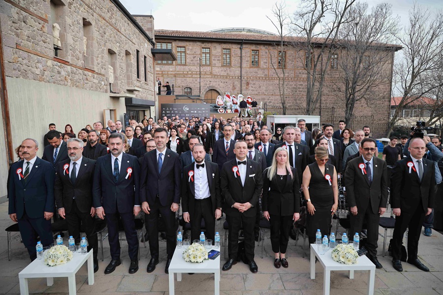 Group of formally dressed people at an outdoor ceremony, standing in front of a brick building with small statues and a crowd behind them, rosettes pinned to their suits and tables with flowers in front.
