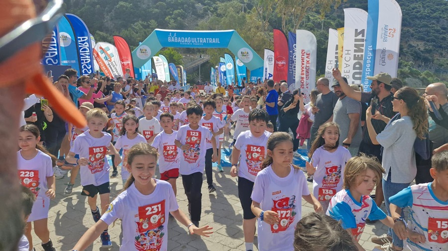 Children's road race: dozens of young runners in numbered shirts sprint toward a blue inflatable arch, while spectators cheer along both sides and sponsor banners flutter above.