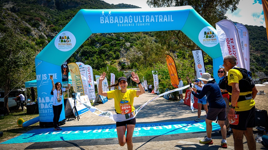 Runner in a yellow shirt crosses the finish line with arms raised as spectators cheer under a blue Babadag Ultra Trail arch in Oludeniz, Turkey.