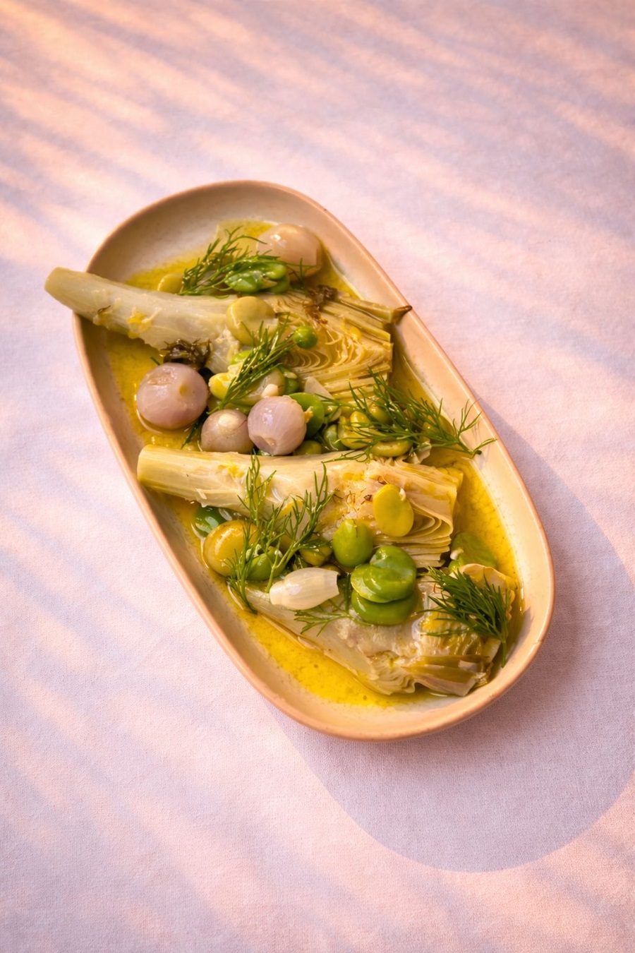 Oval plate with artichoke wedges, peas, pearl onions, and dill in a creamy yellow sauce.