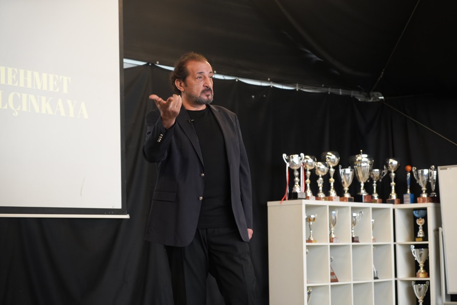 Speaker in a dark blazer gesturing with his right hand on a stage, trophies lined up on shelves behind him