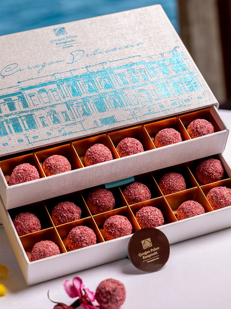 Box of pink sugar-coated chocolate truffles in a white gift box with blue architectural print design, arranged in yellow compartments