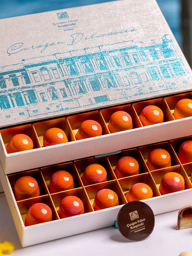 Box of glossy orange confections arranged in a grid; lid features blue illustration of Ciragan Palace Kempinski branding.