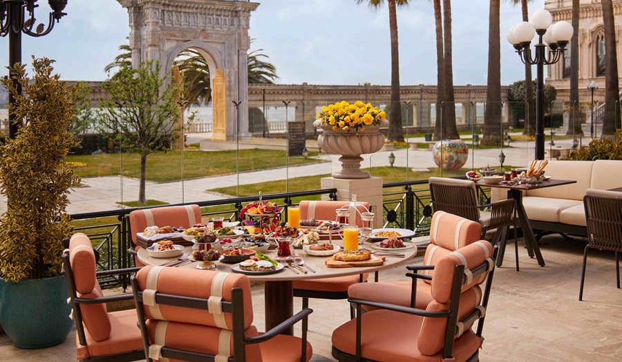 Outdoor terrace breakfast setup: round table with orange cushioned chairs, plates of pastries, fruit, juice, and coffee under a sunny garden with classical arches in the background.