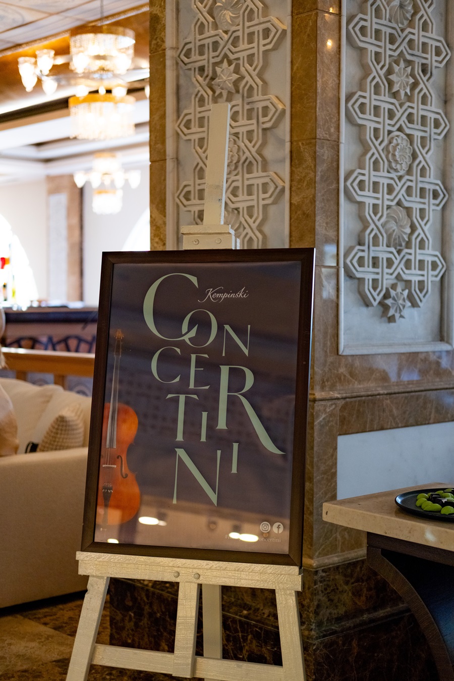 Poster on an easel advertising a Kempinski concert, featuring a violin image and stylized text in a luxurious hotel lobby with marble and ornate wall panels.