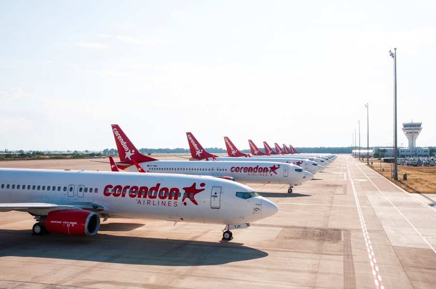 Line of Corendon Airlines jets parked on a sunny airport tarmac, red tails with star logo visible in a row.