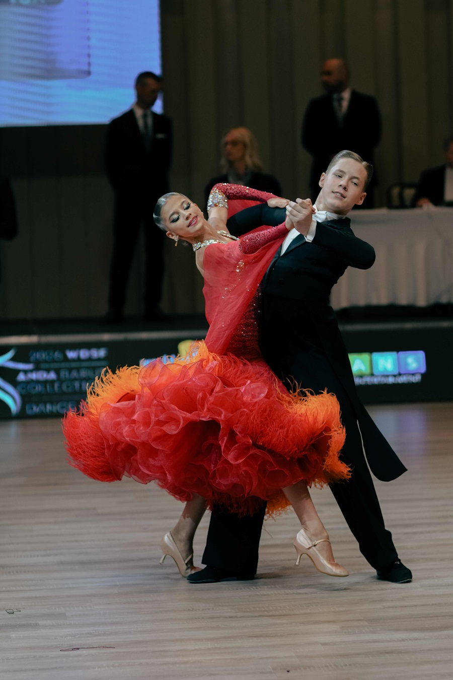 Couple performs a ballroom dance on a wooden floor; woman in a sparkling red gown with ruffles and fringe, man in a black suit.