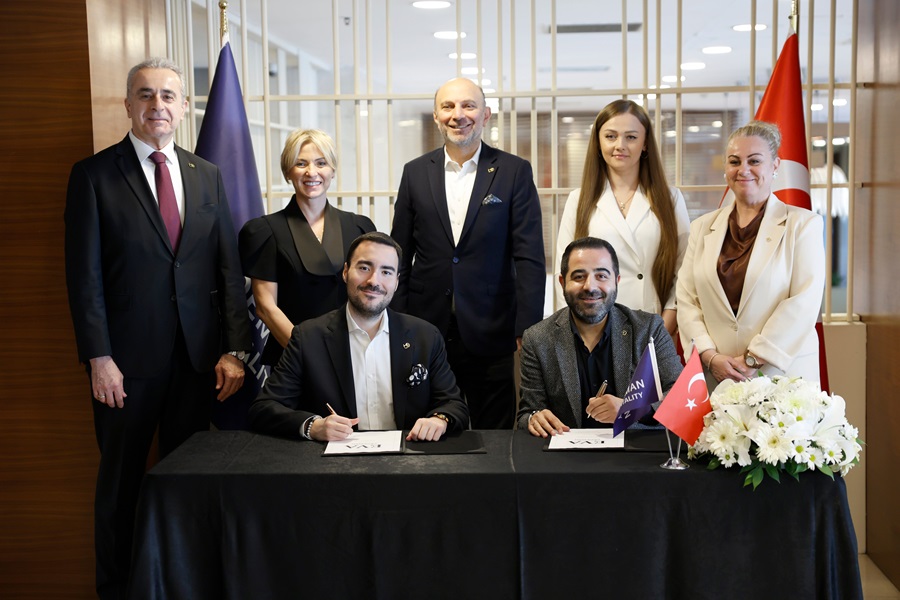 Two men sit at a table signing documents while six colleagues stand behind them in a formal ceremony, with flags and a flower arrangement nearby.
