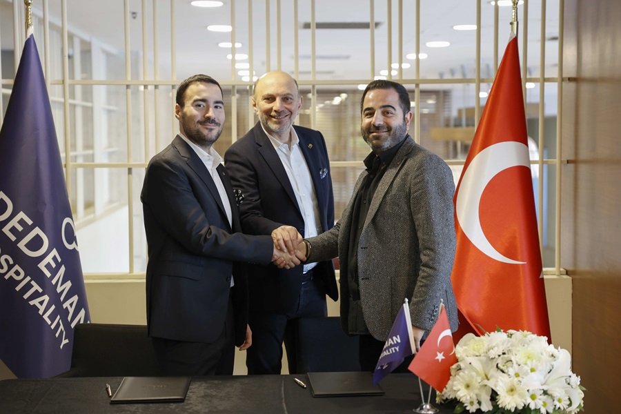 Three men in business suits shake hands in a conference setting, with a Turkish flag and floral arrangement nearby.