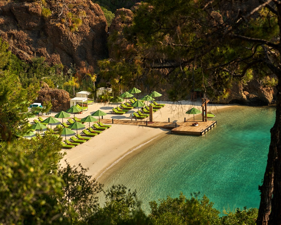Sunny beach with green umbrellas and lounge chairs along a sandy cove, turquoise water, and rocky cliffs nearby.