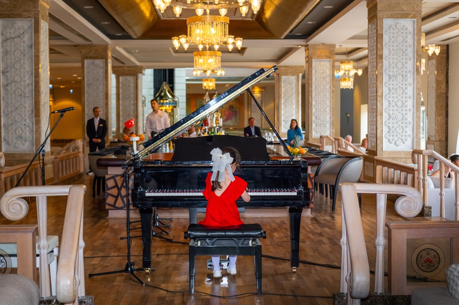 Young girl in a red dress plays a grand piano in an opulent lounge with chandeliers and people in the background.