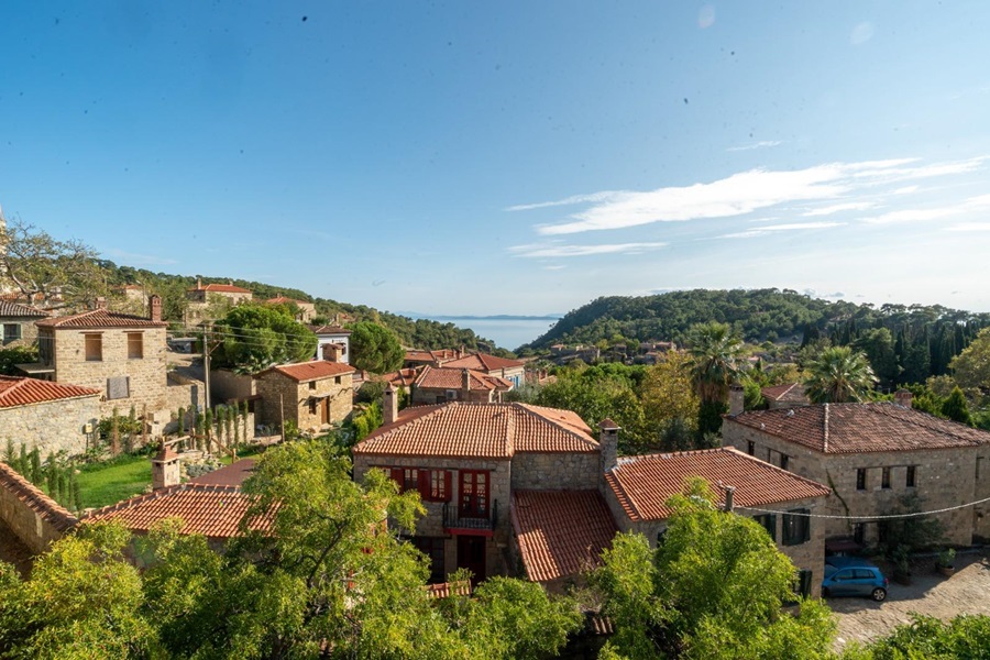 Stone village with red-tiled roofs, green trees, and rolling hills under a blue sky near the coastline