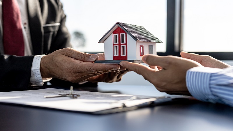 Two professionals exchange a small house model over a tablet, symbolizing a real estate discussion/transaction.