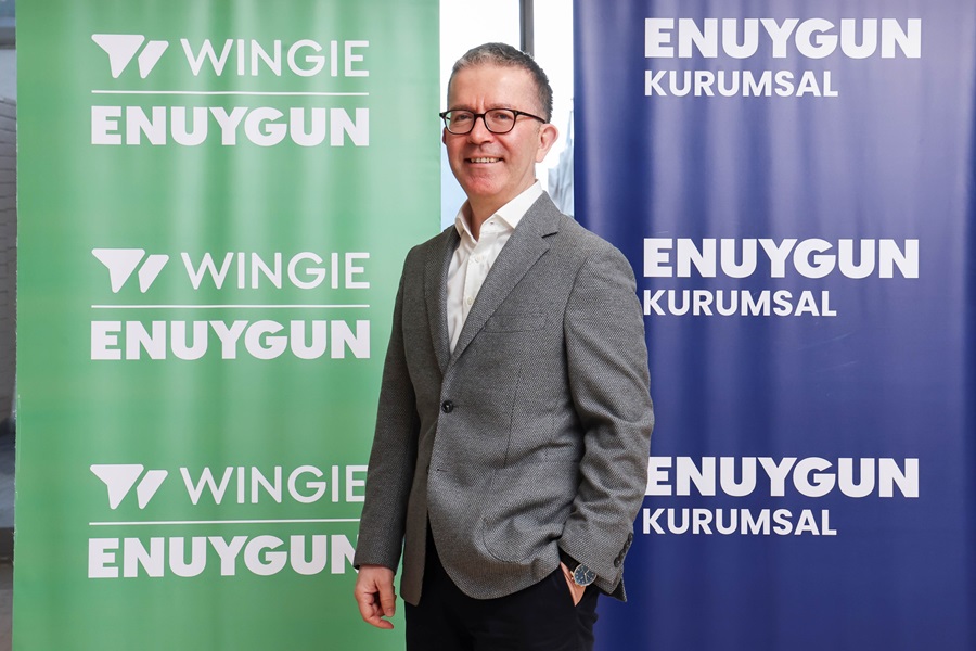 Man in a gray blazer posing between green and blue banners with 'WINGIE ENYUGUN' branding, smiling at camera at a corporate event.
