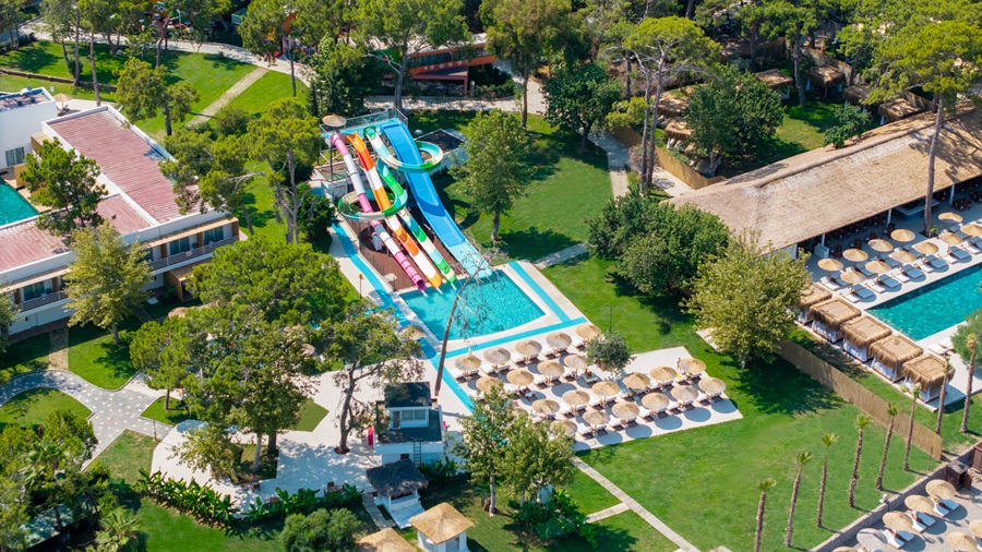 Aerial view of a resort pool area with colorful water slides, a rectangular blue pool, and rows of sun loungers among green lawns and trees.