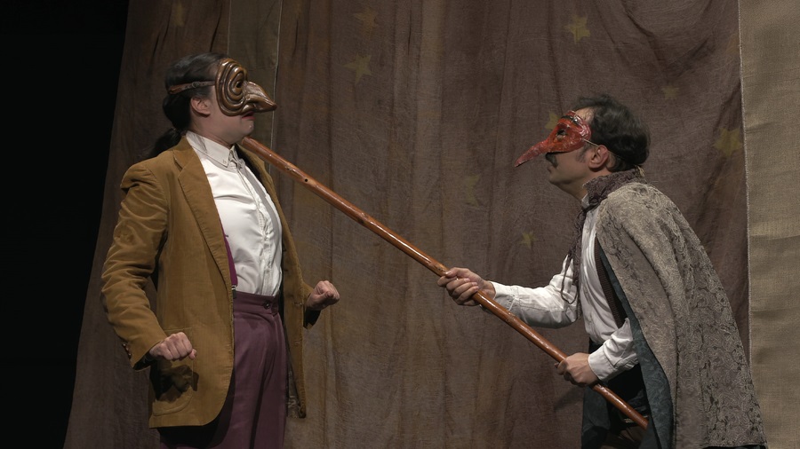 Two masked actors on a theater stage wrestle with a long wooden pole between them, one in a brown blazer and maroon pants and the other in a cloak with a red beaked mask.