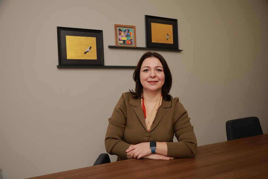 Woman in a brown blazer sits at a wooden conference table, hands folded, smiling in a neutral office setting with framed art on the wall