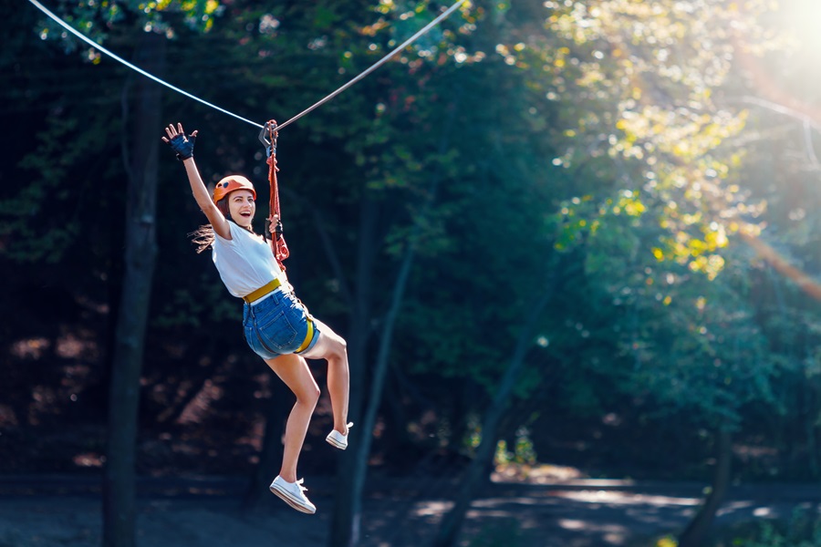 Person zip-lining through a sunlit forest, wearing helmet and harness, arms raised in excitement.
