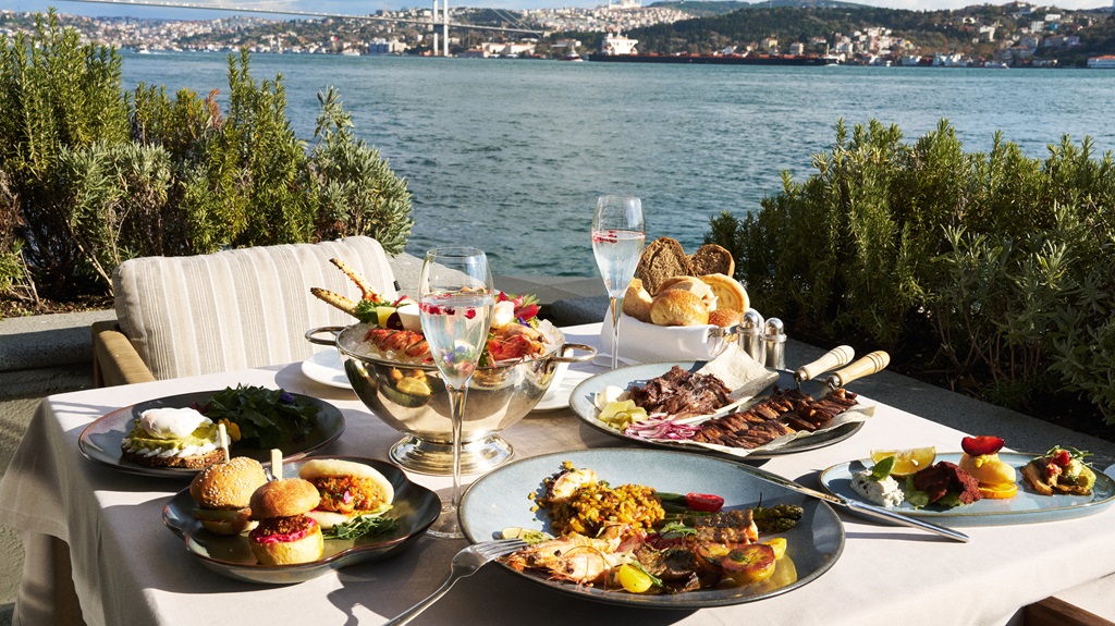 Outdoor coastal dining table with assorted dishes, bread, salads, and champagne by the water with a distant cityscape.