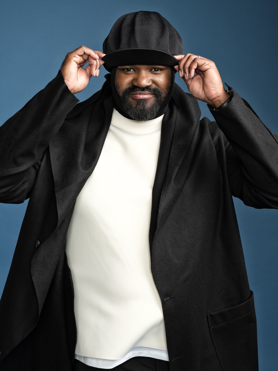 Man in a black coat adjusts his hat while facing the camera against a blue background, smiling subtly.