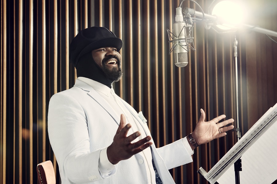 Man in a white suit and black hat sings into a studio microphone in a recording booth, arms open in performance.