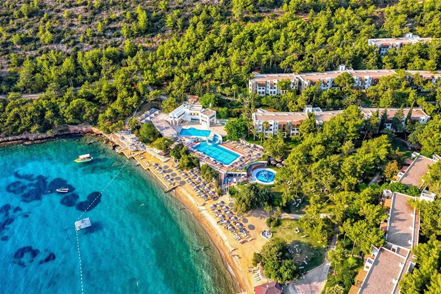 Aerial view of a coastal resort with turquoise water, a sandy beach, sun loungers, and a large pool area surrounded by trees and white buildings.