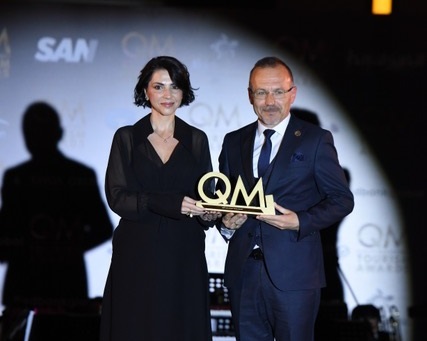 Two well-dressed people on a stage pose for a photo, holding a gold QM award trophy together at an event.