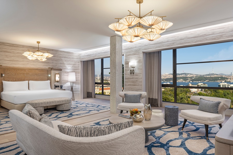 Spacious hotel suite with a seating area, king bed, and large windows overlooking city and water, with modern chandeliers.