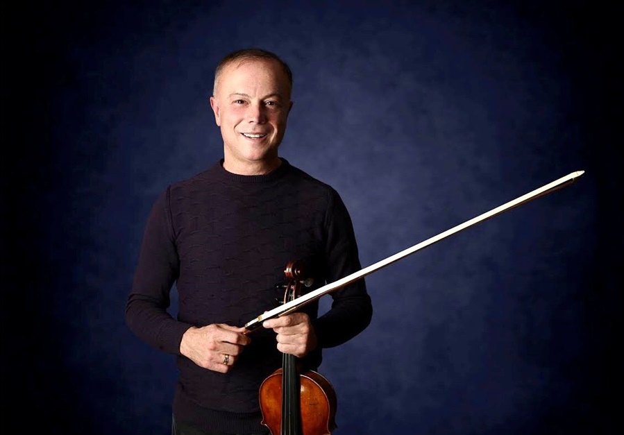 Smiling violinist holds a bow across a violin against a dark blue studio backdrop for a portrait.
