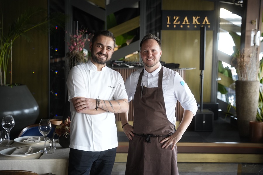 Two chefs pose for a photo in a restaurant; one in a white jacket, the other in a brown apron, smiling at the camera.