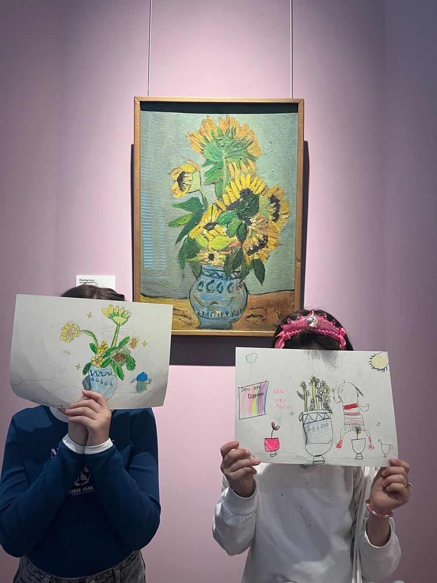 Two children in a gallery hold up drawings of vases with sunflowers in front of a framed sunflower painting.