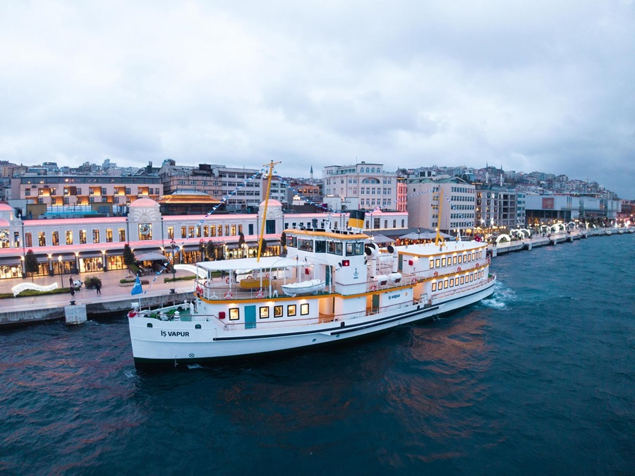 White passenger ferry moored along a lit waterfront promenade with a European city skyline in the background at dusk.