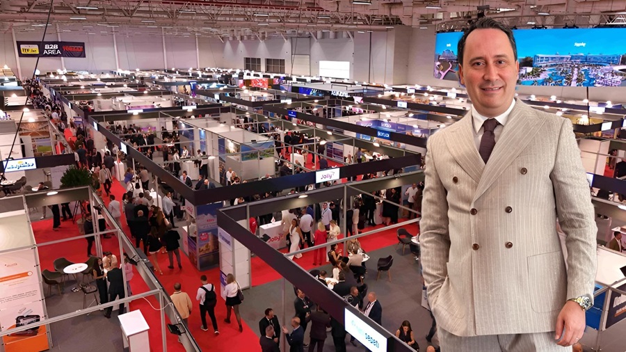 Smiling man in a light suit stands at a busy trade show with numerous booths and attendees in the background.