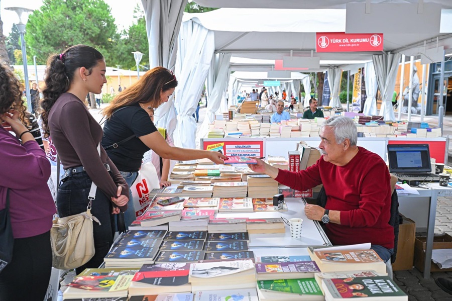 Outdoor book fair: a vendor seated at a table of books hands a book to a young woman customer outdoors under white tents.