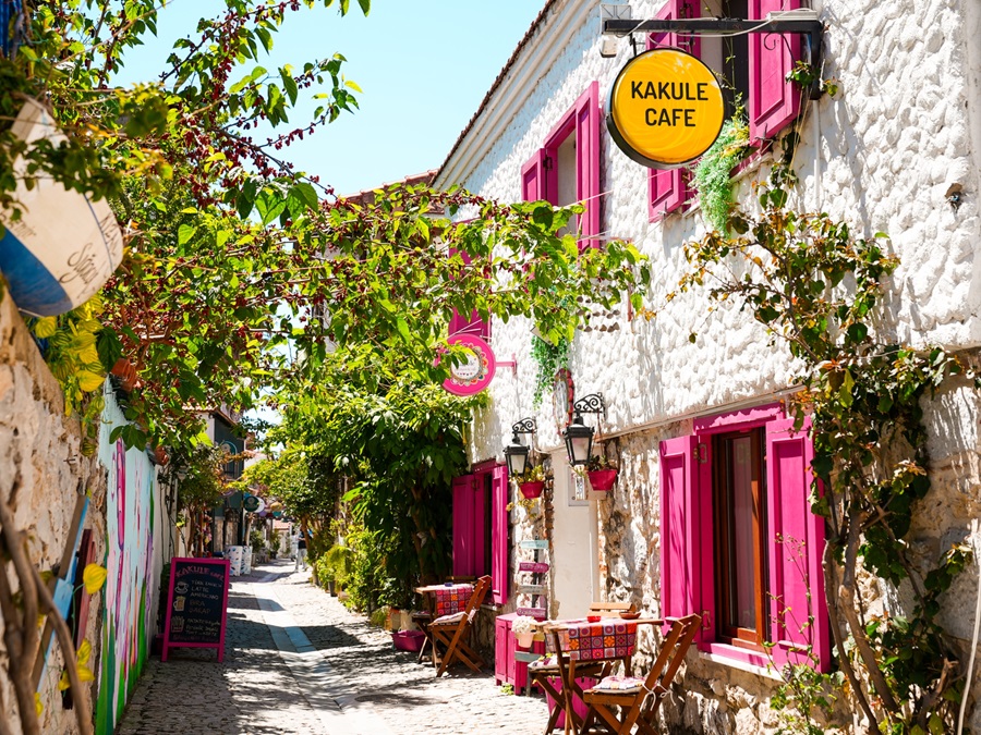 Sunny narrow stone alley with pink window frames, outdoor tables, and a bright yellow Kakule Cafe sign amid green vines.