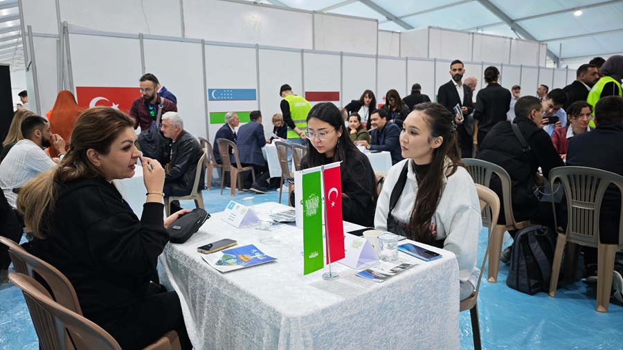 Busy exhibition hall with people seated at tables for discussions; two young women and a woman in a black jacket are talking at a table with flags, Turkish flag visible.