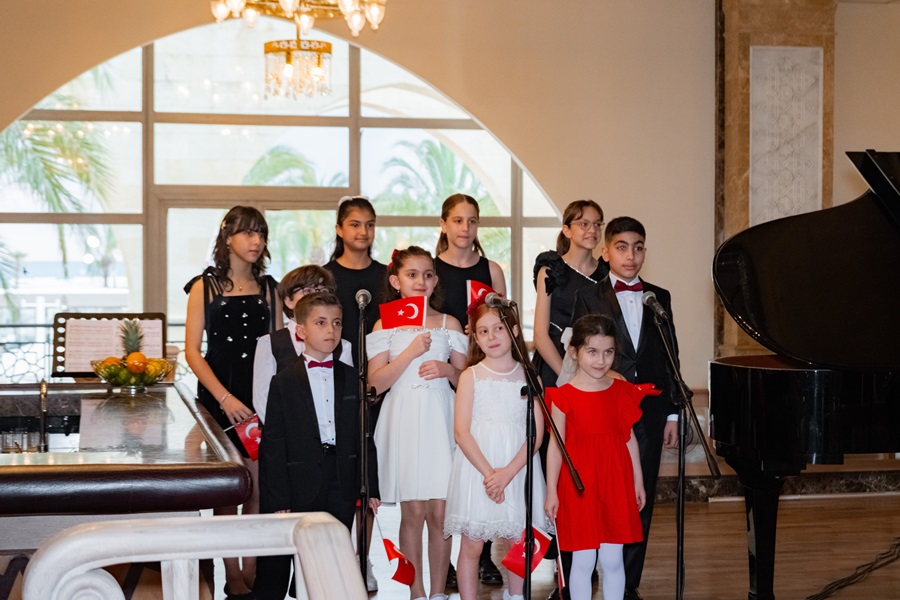 Group of children in formal attire singing into microphones in a room with a grand piano and arched windows, holding Turkish flags.