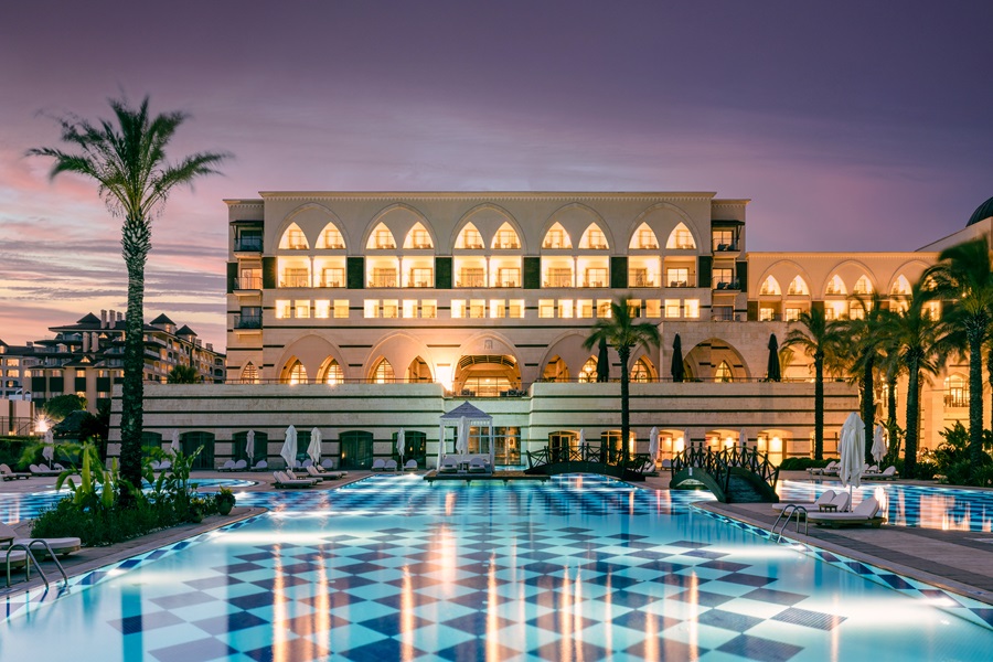 Luxurious resort hotel at dusk with arched windows, palm trees, and a lit swimming pool reflecting the building's lights.