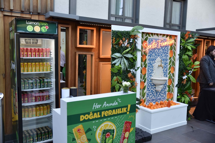 Promotional beverage display with a glass-front fridge of colorful cans next to a green promotional counter outdoors.