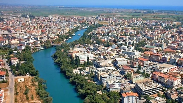 Aerial view of a city with a winding blue river running through dense neighborhoods and tree-lined banks near the coast in the distance.