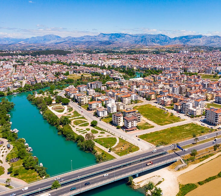 Aerial view of a city with a turquoise river, a bridge, and green parks, set against mountains in the distance.