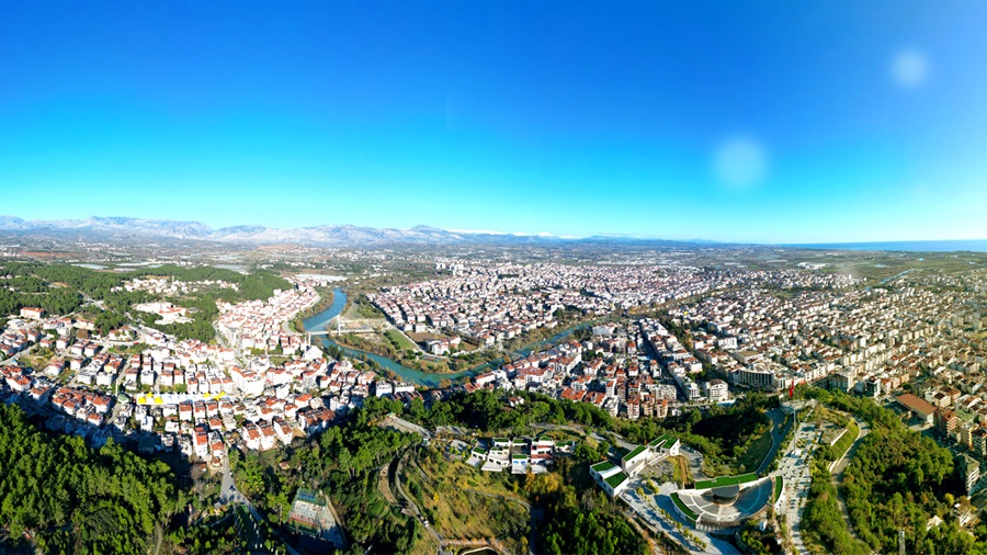 Aerial view of a sprawling city built on hills with a winding river, dense housing, and distant mountains under a clear blue sky.",