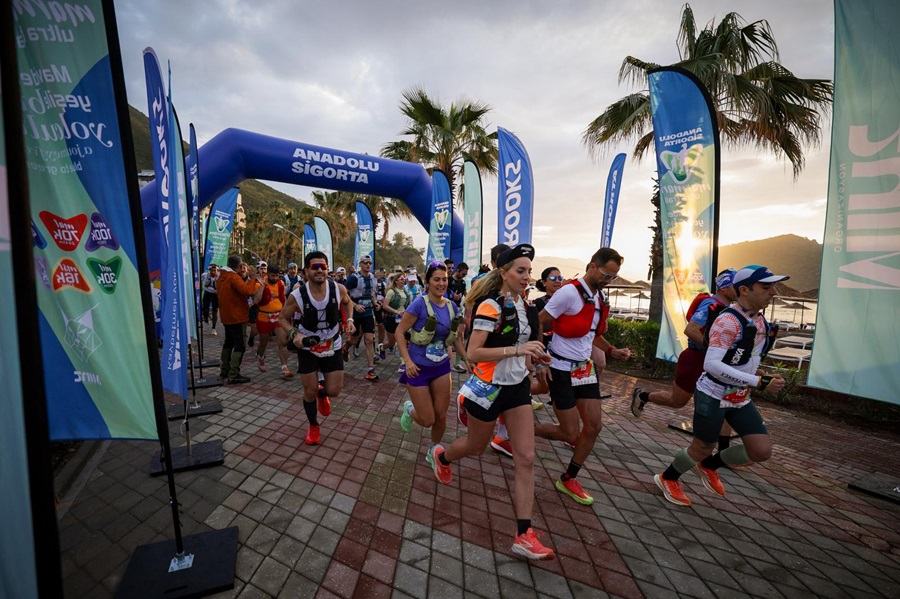 Runners start a race under a blue inflatable arch with Anadolu Sigorta branding, flanked by colorful banners and palm trees at sunset.